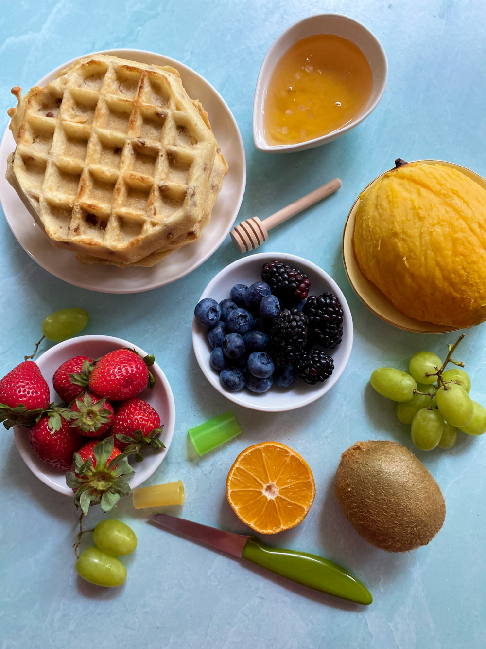 a flat lay showing waffles in a plate, honey in a bowl and several types of fruits