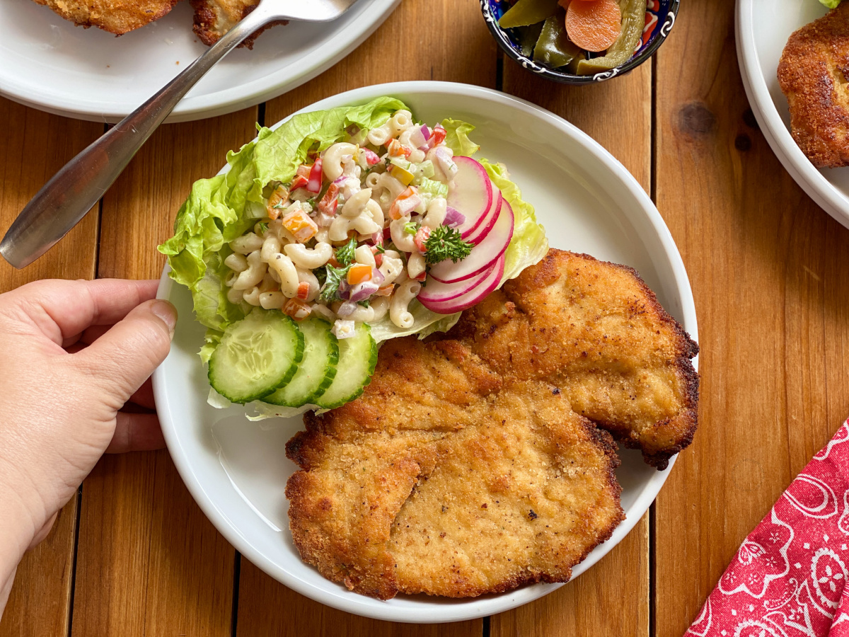 a hand holding a plate with a chicken milanesa and macaroni salad