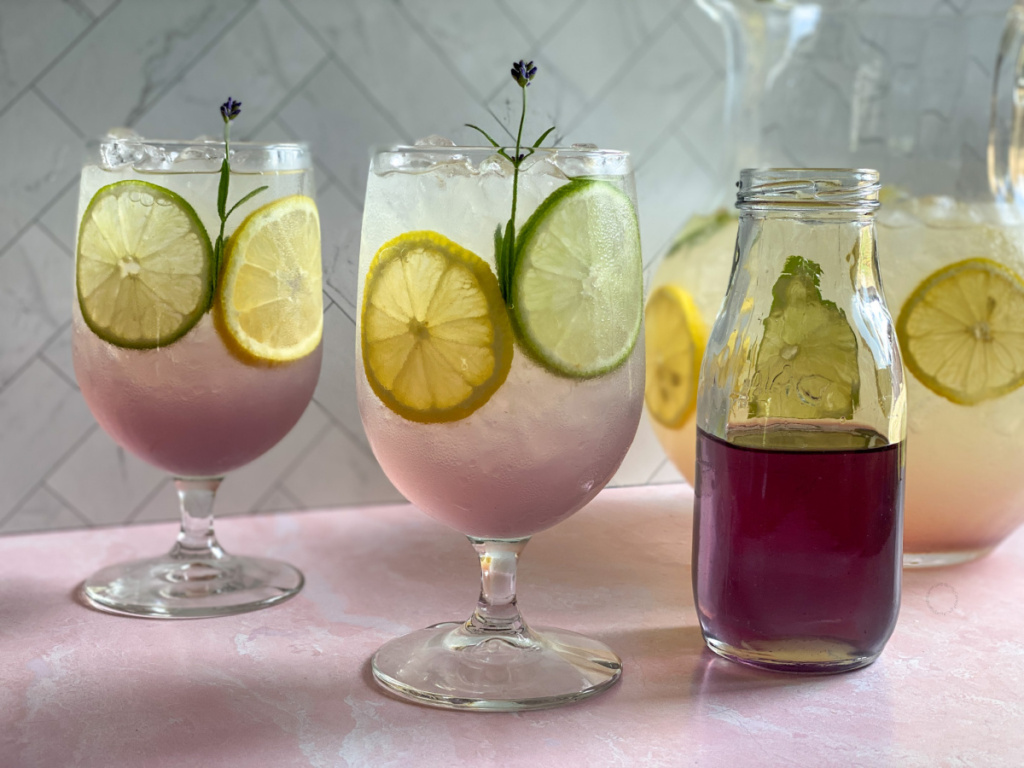 Two glasses with lavender lemonade and a bottle with culinary lavender and blue pea flower syrup