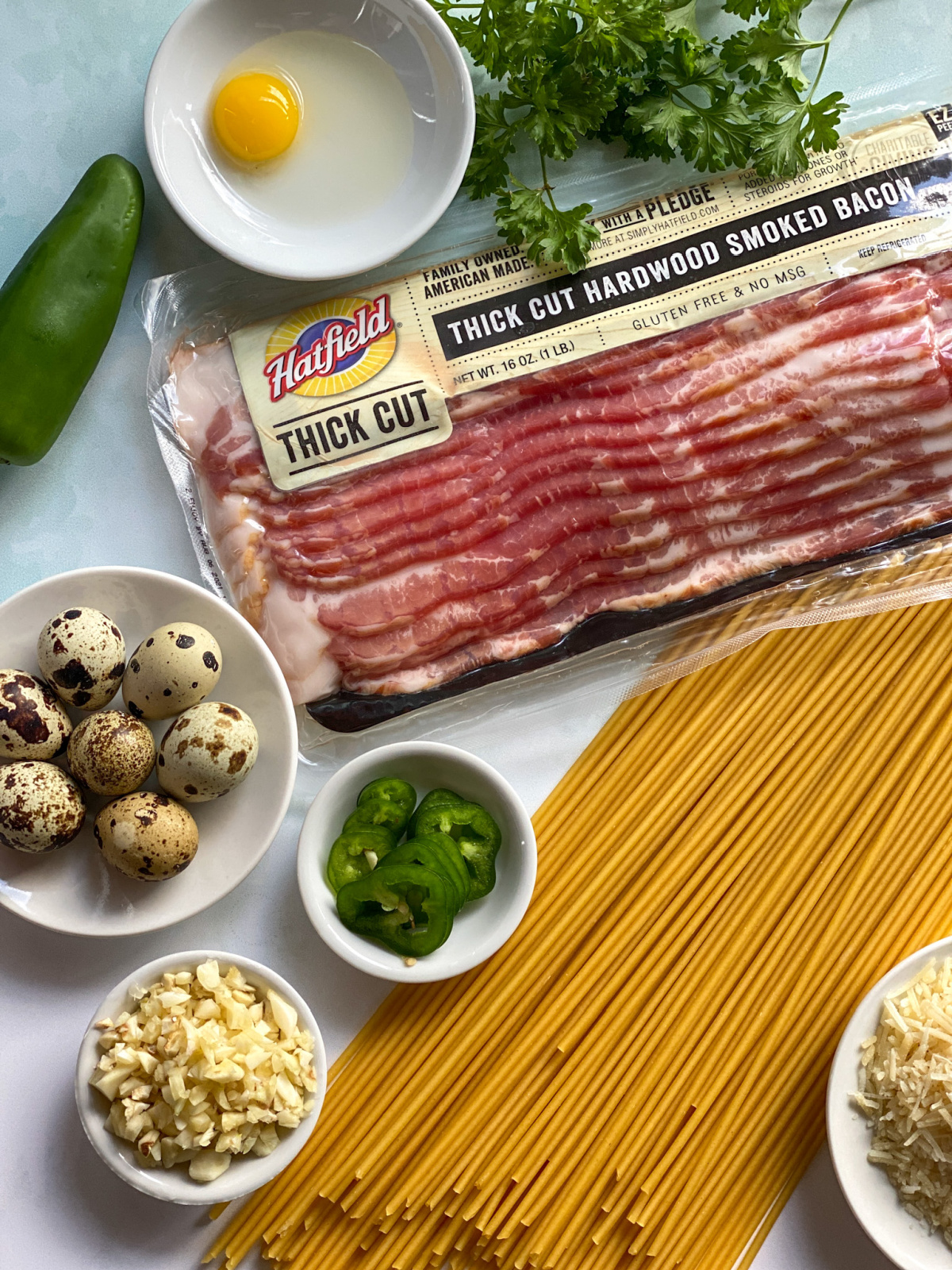 A top shot showing pasta, bacon, chopped garlic, jalapeño pepper, and fresh parsley