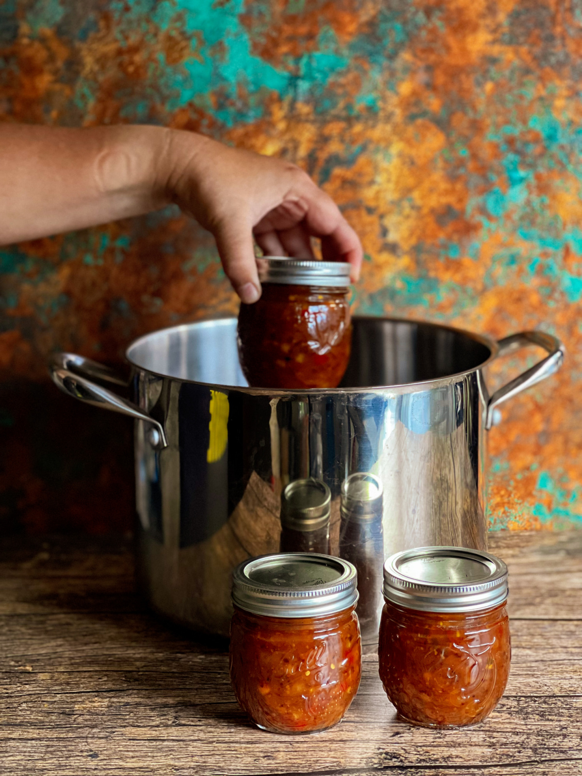 a hand introducing a mason jar into a canning pot