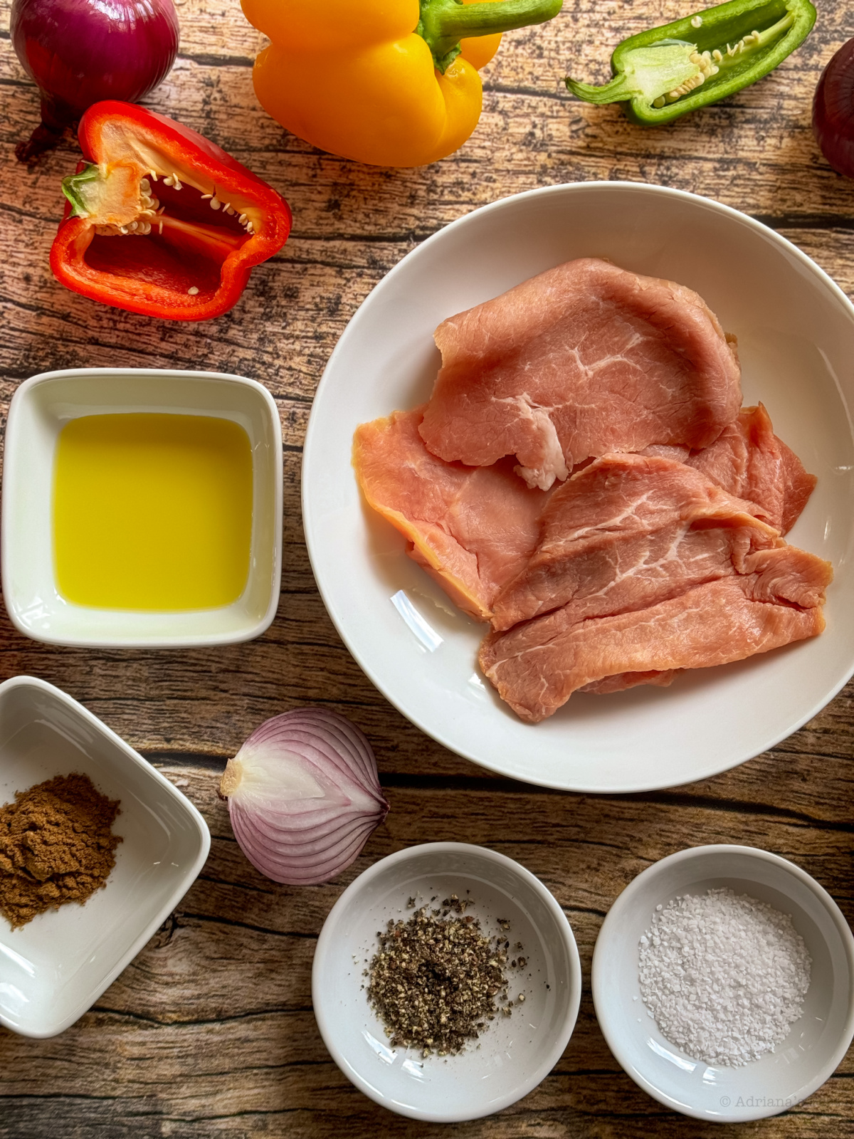 Una superficie de madera con varios ingredientes: escalopes de ternera cruda en un plato, aceite de oliva, especias, sal, pimienta, cebolla y pimientos en rodajas de color amarillo, rojo y verde.