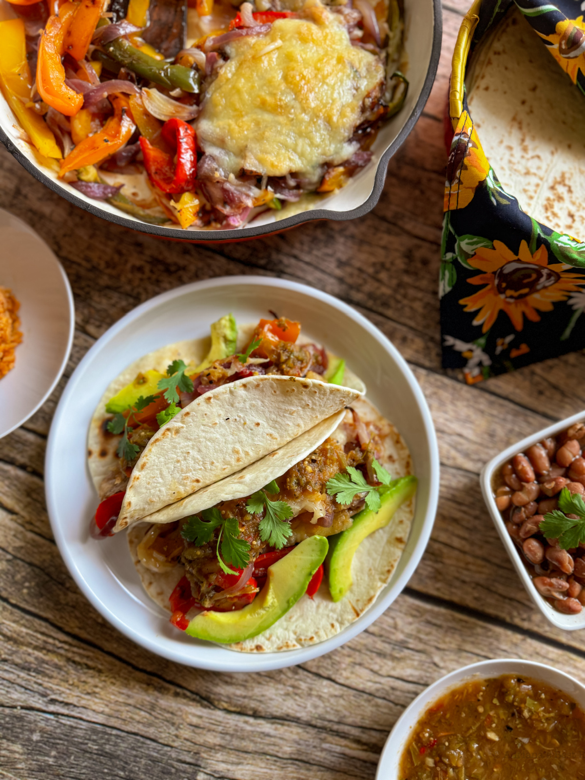 Un plato con dos tacos rellenos de fajitas de ternera asada estilo Tex-Mex, vegetales mixtos, rodajas de aguacate y cilantro se encuentra sobre una mesa de madera, rodeado de platos de frijoles, y salsa 